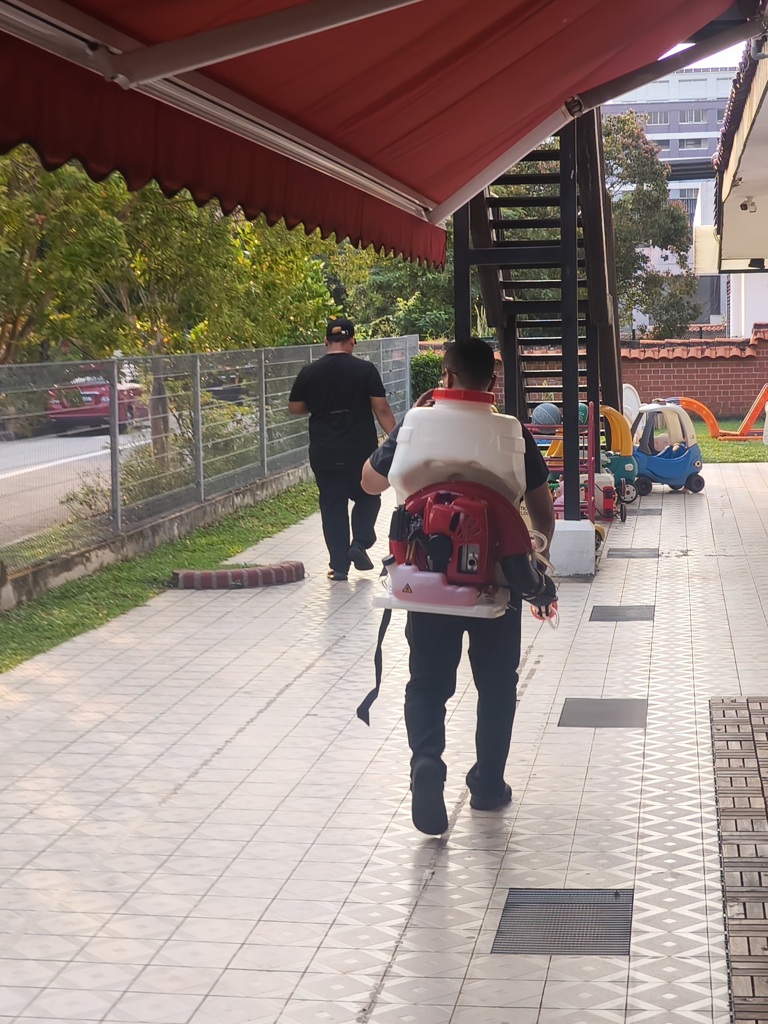 Professional pest control technician spraying outdoor area at MapleBear Jurong West, children's play area visible in background